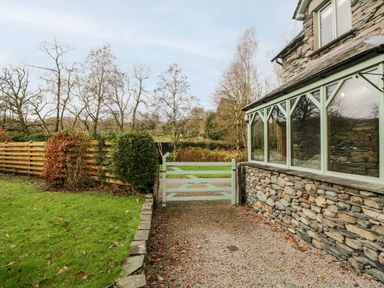 A garden area with a gravel path green grass a wooden fence and a stone house with large windows at Scandale Bridge Cottage in Ambleside