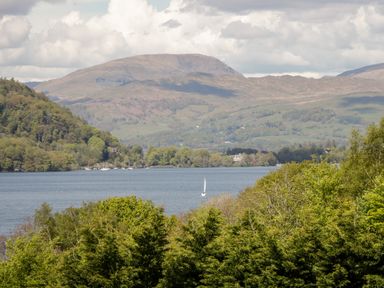 A lake surrounded by trees with mountains in the background and a sailboat on the water at Scandale Bridge Cottage in Ambleside