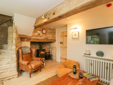 A living room with a leather armchair a fireplace with a wood burning stove a wooden door and a mounted TV at Thorn Cottage in Fairford