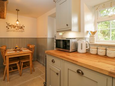 A kitchen corner with wooden table and chairs next to cabinets with microwave kettle and containers at Thorn Cottage in Fairford