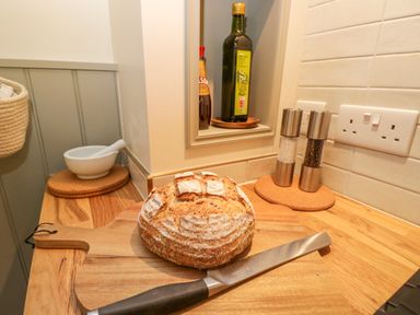 A kitchen countertop with a loaf of bread on a wooden cutting board a bread knife a mortar and pestle oil and sauce bottles salt and pepper grinders at Thorn Cottage in Fairford