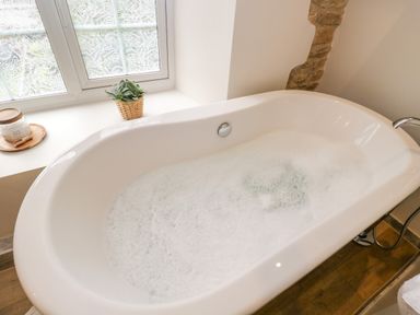 A bathroom with a freestanding bathtub filled with soapy water and a window ledge with a plant and bath salts at Thorn Cottage in Fairford