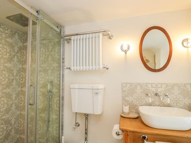 A bathroom with a shower enclosure tiled with patterned tiles a white toilet a wooden vanity with a white sink an oval mirror and wall sconces at Thorn Cottage in Fairford