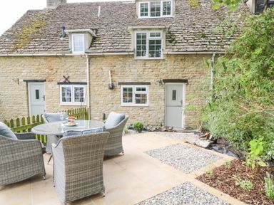 An outdoor patio area with a round glass table and four wicker chairs in front of a stone cottage with multiple windows and doors surrounded by greenery at Thorn Cottage in Fairford