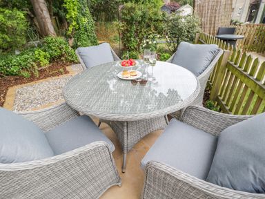 An outdoor seating area with a round glass-topped wicker table four cushioned wicker chairs plates of strawberries and scones glasses of drink and a pair of sunglasses in a garden at Thorn Cottage in Fairford