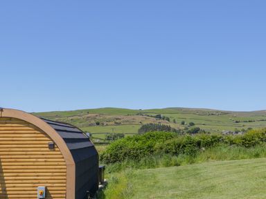 A curved wooden cabin beside a grassy field with green hills under a clear blue sky at Holly in Ulverston