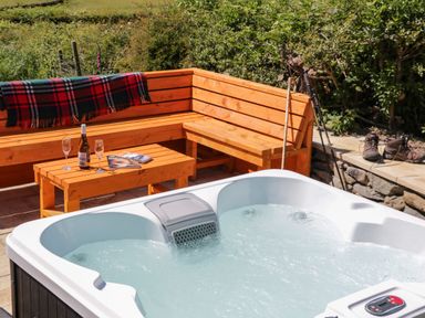 An outdoor seating area with wooden benches a table with a bottle and glasses and a hot tub at Holly in Ulverston