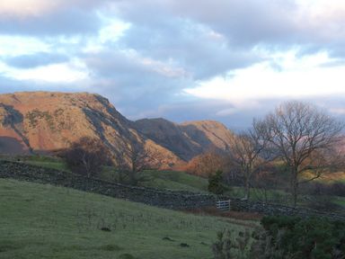 A landscape with grassy fields trees a stone wall wooden gate and rocky hills under a cloudy sky at Holly in Ulverston