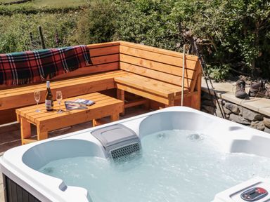 An outdoor hot tub with bubbling water next to a wooden bench and table with a bottle of wine glasses and magazines Rowan Ulverston