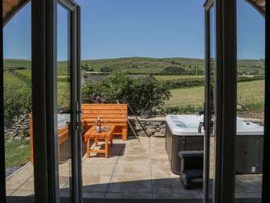 A patio with a wooden bench and table set and a hot tub with steps in a countryside setting at Rowan in Ulverston
