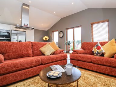 A living room with two red sofas a round coffee table with mugs and pastries a kitchen area and windows with blinds at Woodbridge Penthouse in Woodbridge
