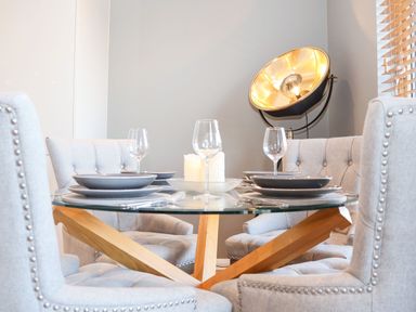 A dining area with a glass table set with plates and wine glasses surrounded by upholstered chairs and a large floor lamp at Woodbridge Penthouse in Woodbridge