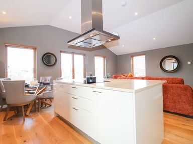 An open plan kitchen and living area with a white island unit wooden floor grey walls dining table with chairs and an orange sofa at Woodbridge Penthouse in Woodbridge