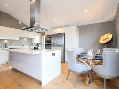 A kitchen with a white island and stainless steel appliances next to a dining area with a glass table and four upholstered chairs at Woodbridge Penthouse in Woodbridge