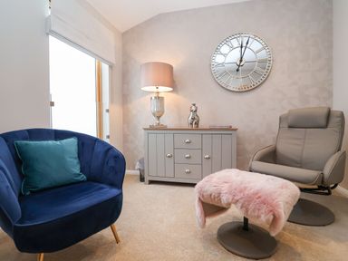 A room with a blue armchair with a green pillow a gray armchair with a pink fur-covered footrest a gray sideboard with a lamp and decor and a large wall clock at Woodbridge Penthouse in Woodbridge