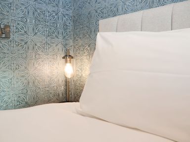A bed with white pillows and headboard next to a bedside lamp with patterned wallpaper at Woodbridge Penthouse in Woodbridge