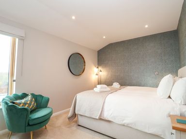A bedroom with a double bed, round wall mirror, green armchair with cushion, bedside table, and patterned wallpaper at Woodbridge Penthouse in Woodbridge