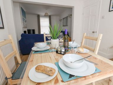A dining table set with white plates bowls bread and a bottle of wine in a room with wooden chairs and a blue sofa at Day Cottage in Weymouth