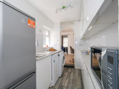 A narrow kitchen with a refrigerator, washing machine, microwave, sink, and countertop leading to an adjoining living room at Day Cottage in Weymouth