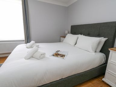 A bedroom with a double bed with white linens and pillows three rolled white towels and an open magazine on the bed two nightstands with lamps and a window with curtains at Day Cottage Weymouth