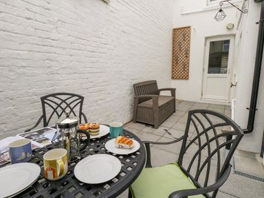 A small outdoor patio with a table set with cups, plates, and pastries and a bench against a white brick wall at Day Cottage in Weymouth