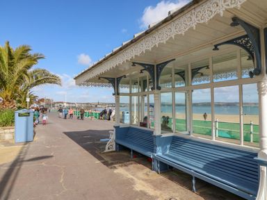 A covered blue bench with decorative white trim on a seaside promenade with people walking and a beach in the background at Day Cottage in Weymouth