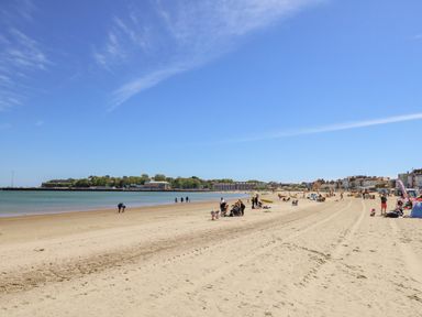 A sandy beach with people walking and sitting near the water and buildings in the background at Day Cottage Weymouth