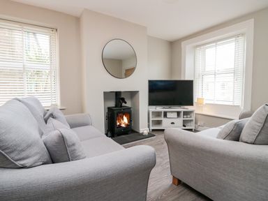 A living room with two grey sofas a wood-burning stove a round mirror a television on a white stand and windows with blinds at Wainwright Cottage in Gosforth