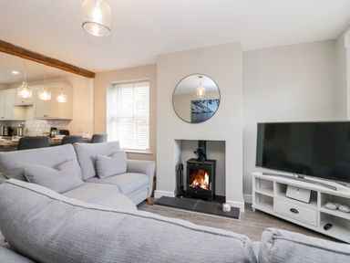 A living room with gray sofas a wood stove a round mirror a television and an open view into a kitchen at Wainwright Cottage in Gosforth