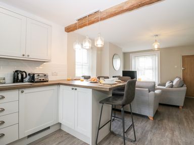 A kitchen with white cabinets and wooden countertop next to a living room with gray sofas at Wainwright Cottage in Gosforth