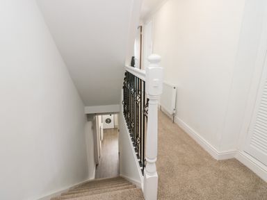 A stairway with beige carpet and white walls leading down to a hallway with wooden flooring and a washing machine visible at the end at Wainwright Cottage in Gosforth