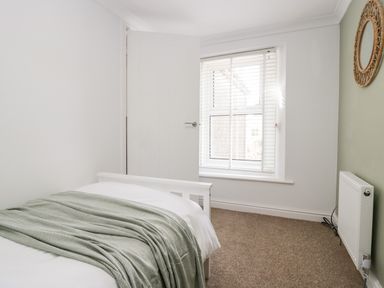 A bedroom with a bed covered with a green blanket and white sheets near a window with blinds and a round woven mirror on the wall at Wainwright Cottage in Gosforth