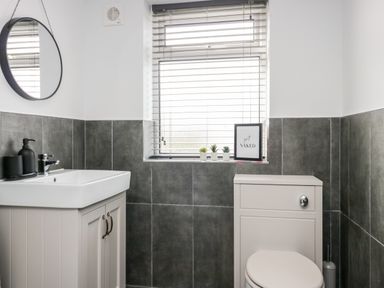 A bathroom with a sink vanity, round mirror, toilet, window with blinds, small plants, and a framed sign at Wainwright Cottage in Gosforth