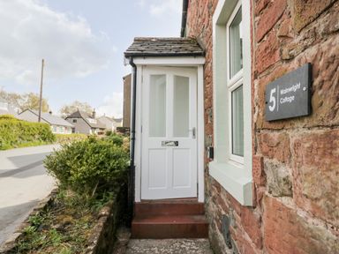 A white door entrance with a mail slot and steps on a stone building with a number 5 Wainwright Cottage sign in Gosforth