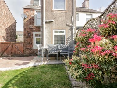 A backyard with a patio table and chairs near a house and flowering bushes at Wainwright Cottage in Gosforth