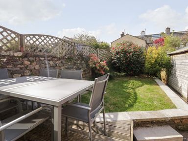 An outdoor garden area with a table and chairs on a deck and grass with bushes and a stone wall at Wainwright Cottage in Gosforth