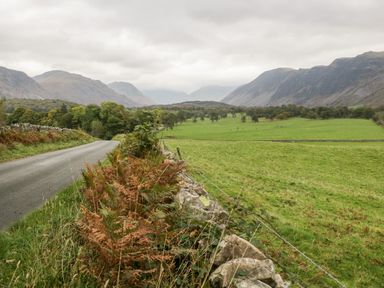 A countryside road beside a stone wall and green fields with mountains in the background at Wainwright Cottage in Gosforth