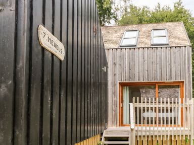 A wooden house with large windows and two roof skylights next to a black wooden wall with a sign reading 7 Meads at Lodge 6 in Corfe Castle