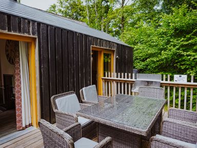 An outdoor deck with a glass-top table and six wicker chairs next to a black wood-paneled building at Lodge 6 in Corfe Castle