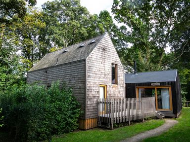 A wooden house with a deck and a smaller black building next to it surrounded by trees at Lodge 11 in Corfe Castle