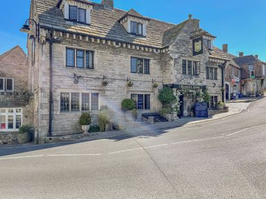 A stone building with multiple windows and potted plants outside on a street corner with a sign reading Bankes Arms in Corfe Castle