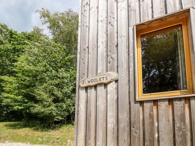 A wooden exterior wall with a window and a wooden sign that reads 21 Wooletts surrounded by green trees at Lodge 21 in Corfe Castle