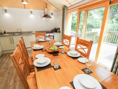 A dining area with wooden table and chairs set with dishes and glasses next to a kitchen and sliding glass doors at Lodge 21 in Corfe Castle