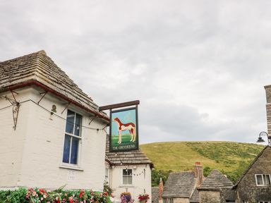 A street view with a building featuring a hanging sign of a greyhound dog and flowers below the window at Lodge 21 in Corfe Castle