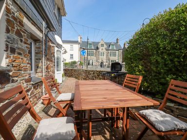 An outdoor patio area with a wooden table, four cushioned wooden chairs, a barbecue grill, stone walls, and a bush at Falcon House in Tintagel