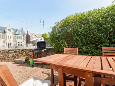 An outdoor patio with a wooden table and chairs a barbecue grill and a stone wall with buildings in the background at Falcon House in Tintagel