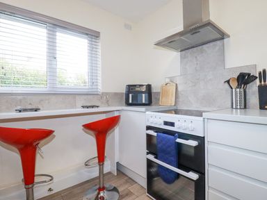 A kitchen with two red bar stools a stove with a blue oven mitt a black air fryer wooden utensils a knife block and a window with blinds at Falcon House in Tintagel