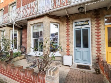 An exterior view of a brick townhouse with a blue front door labeled ELKHORN and a small front garden with plants in Southwold