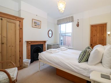 A bedroom with a bed with white linens and striped pillows a wooden wardrobe a fireplace a wooden chair a window with a striped blind and a wooden door at Elkhorn Southwold