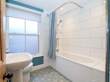 A bathroom with a bathtub shower combination a white sink and toilet a window with a blind and tiled floor at Elkhorn in Southwold
